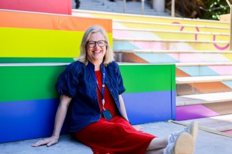 A person sits on the ground leaning against brightly coloured tiered seating, wearing a dress, sneakers, and a lanyard, with rainbow‑coloured steps behind.
