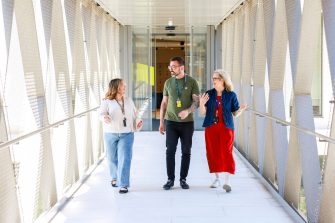 Three people wearing lanyards walk side by side through a bright covered walkway, talking as sunlight filters through metal mesh walls toward glass doors ahead.