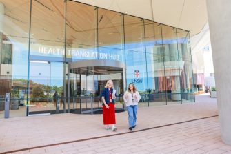 Two people walk away from the glass entrance of the Health Translation Hub at UNSW Sydney, passing through a wide paved forecourt with campus buildings behind.