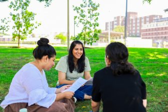 Three people sit on grass in a park-like campus setting, facing each other in a small circle, with one person holding printed papers and trees and buildings in the background.