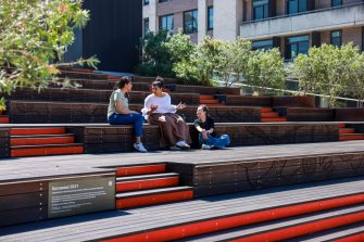 Three people sit spaced along tiered wooden outdoor seating, talking together in a landscaped courtyard with trees and a multi‑storey building in the background.