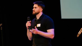 A man speaks into a microphone while presenting on stage at an event.
