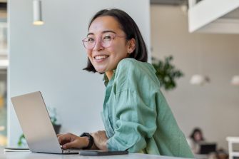 Pretty cheerful Asian woman in eyeglasses and casual clothes browses laptop computer connected to 4g internet updates software uses modern technologies poses in cafeteria looks gladfully away