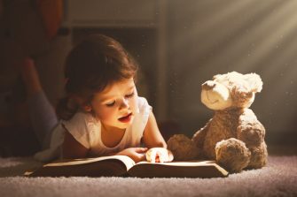 A child reading a book in evening in dark with a toy bear