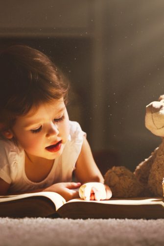 A child reading a book in evening in dark with a toy bear