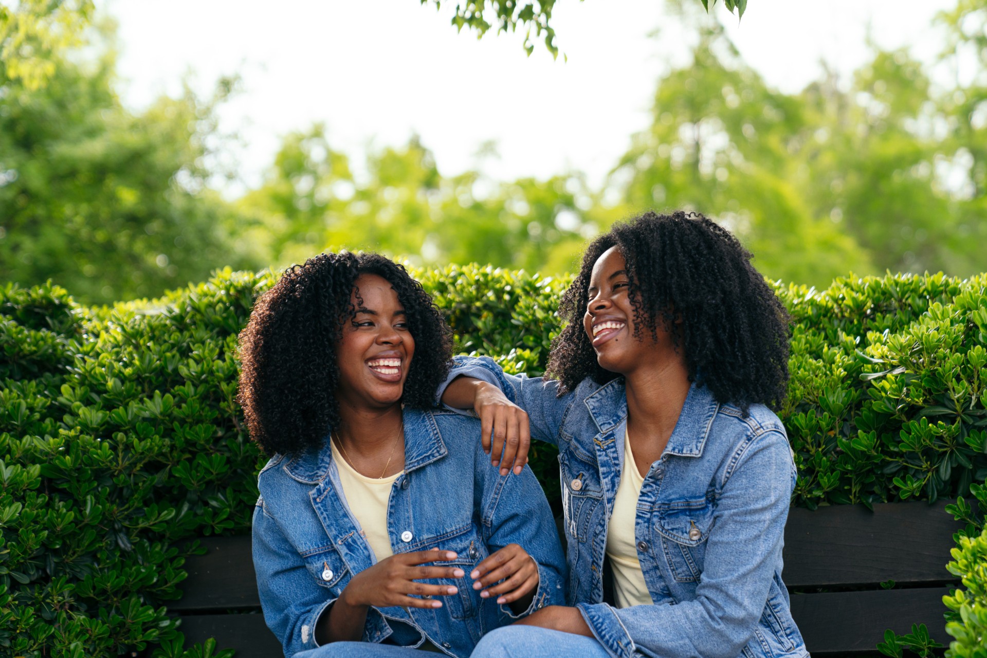 two happy twin sisters are sitting on a park bench, wearing denim jackets and laughing together on a sunny day