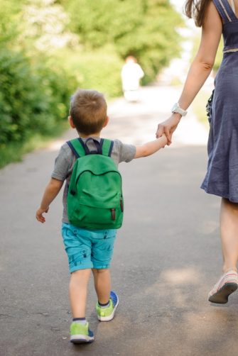 Back view of mother walking down the street with a little son with a backpack on sunny summer day