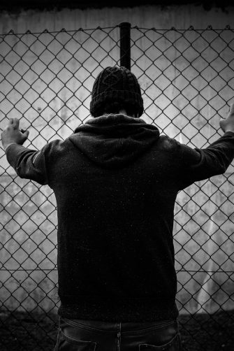A black and white shot from behind of a young male detainee  in front of a fence