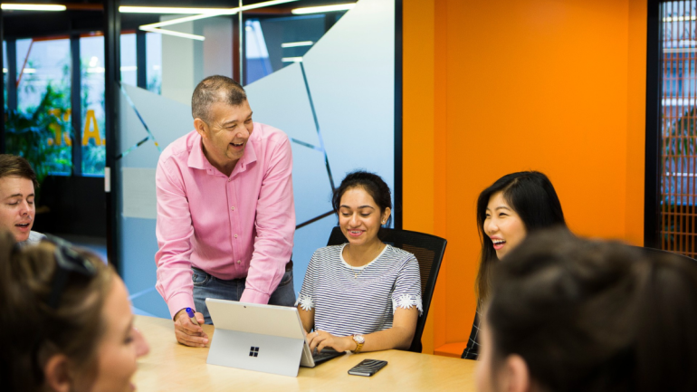 A UNSW lecturer in a pink shirt stands smiling beside students in a bright classroom, as they collaborate around a table with laptops. The scene reflects inclusive, joyful learning central to Universal Design for Learning 3.0.
