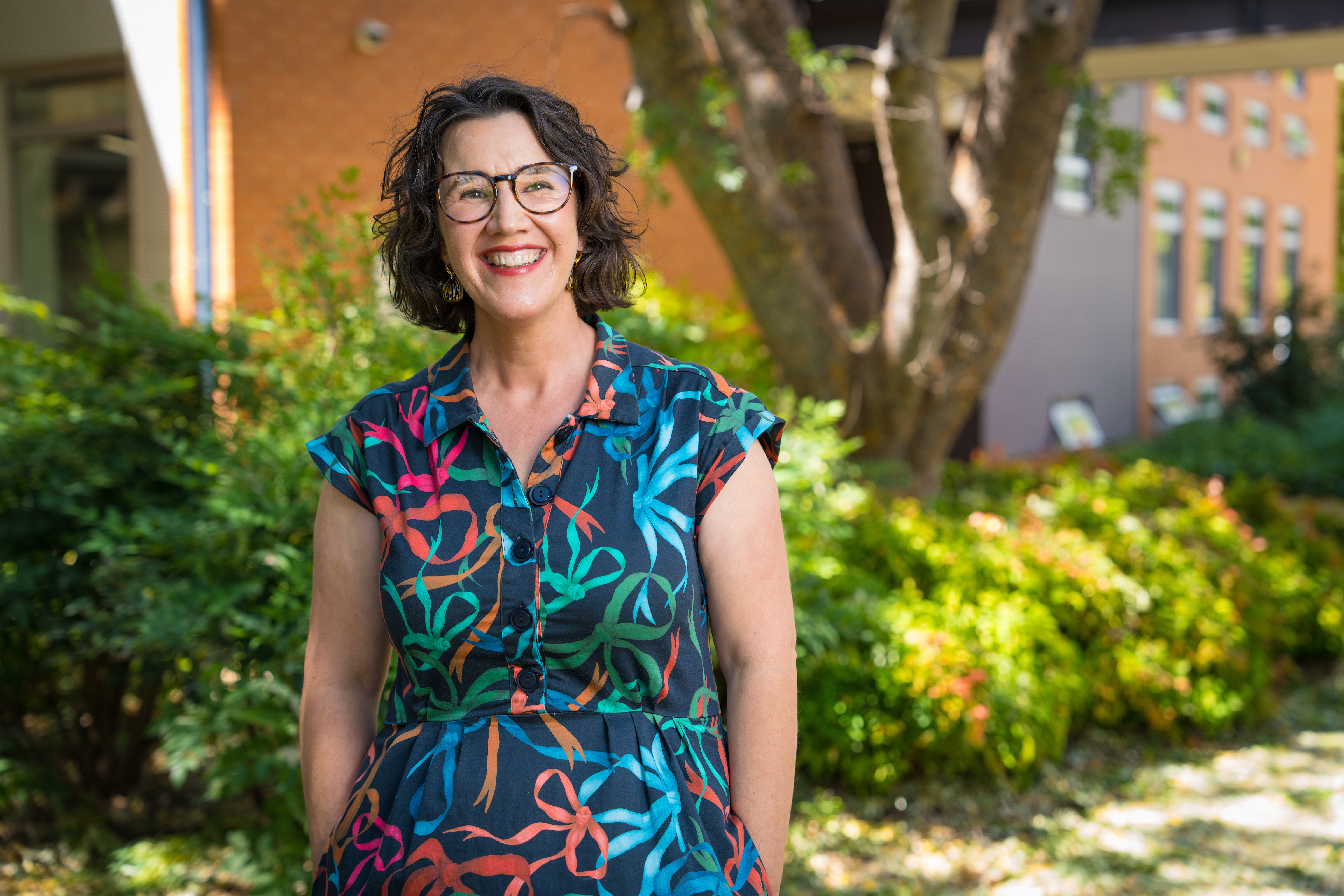 A woman wearing glasses standing and smiling.