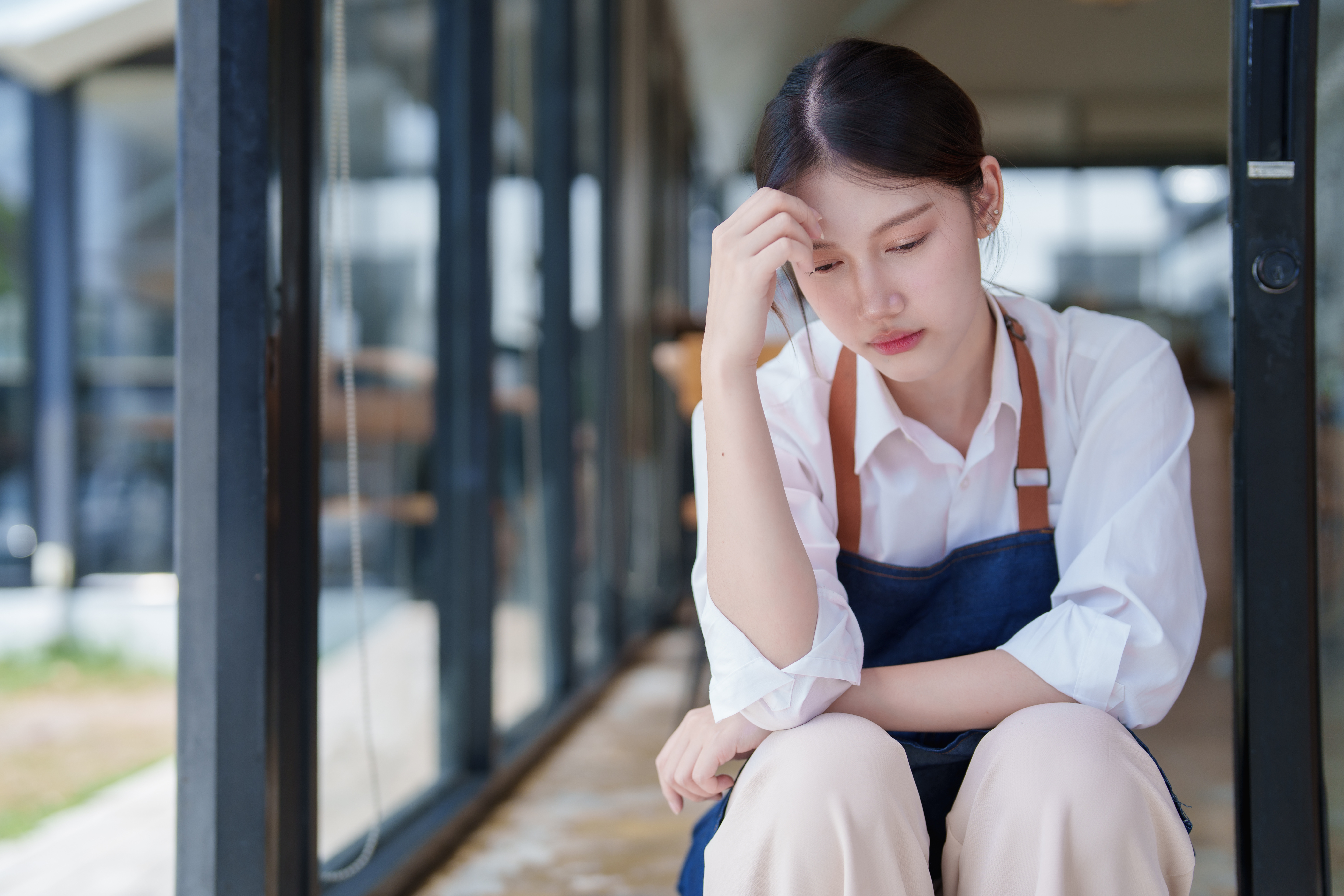 Young woman in apron sitting outside coffee shop feeling overwhelmed and frustrated