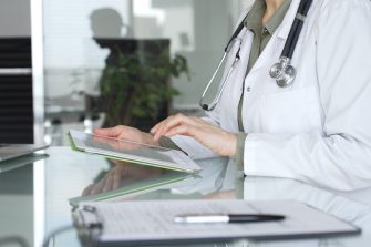 Doctor in white coat with stethoscope using tablet at office desk, reviewing digital patient data for telehealth and clinical care innovations. Medicine and health care concept