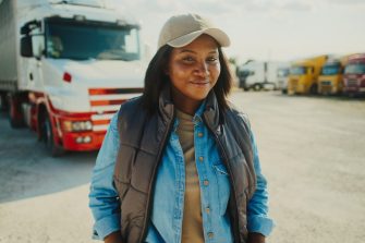 Woman standing in front of truck parking lot. Looking at camera while smiling. Resting in fresh air. Clear sky with sun shining bright. Morning before trucker departs to location