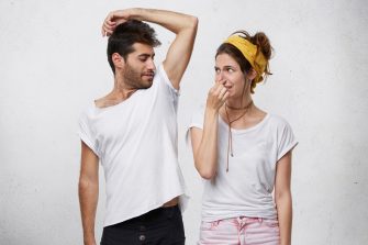 Female having disgusted look covering her nose with hand while smelling her husband`s armpit. Bearded man having nasty smell from sweat after running showing his wet underarm. Unpleasant smell