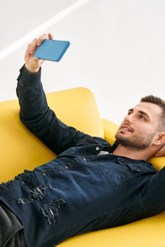 Relaxed man lying on yellow sofa talk on video call by his mobile phone over white background. Social media concept