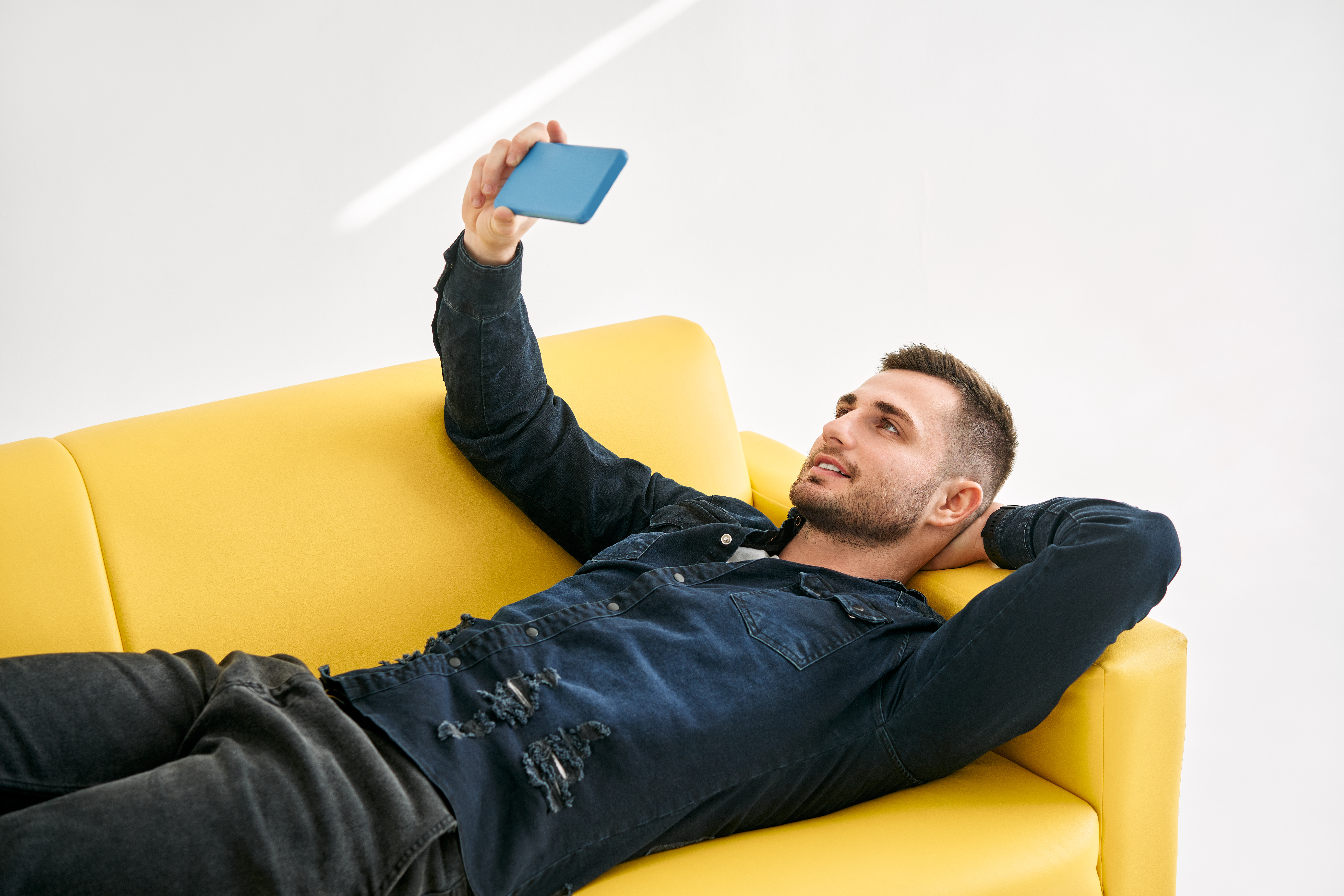 Relaxed man lying on yellow sofa talk on video call by his mobile phone over white background. Social media concept