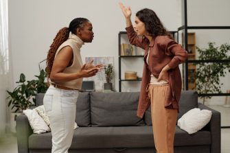 Side view of two young angry girlfriends shouting at one another and gesturing by hands while standing by couch during argument