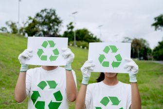 Two volunteers holding recycle signs covering faces, wearing eco friendly t-shirts in park. Environmental conservation campaign, green lifestyle concept, global teamwork for climate awareness.