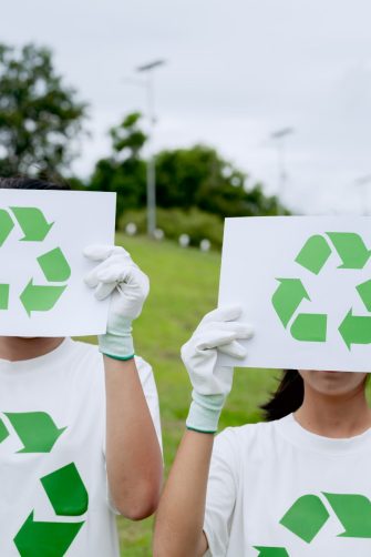 Two volunteers holding recycle signs covering faces, wearing eco friendly t-shirts in park. Environmental conservation campaign, green lifestyle concept, global teamwork for climate awareness.