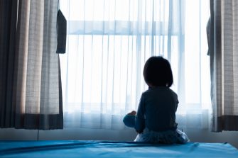 Little baby child sitting on bed playing with the doll in the dark room with light throught curtain
