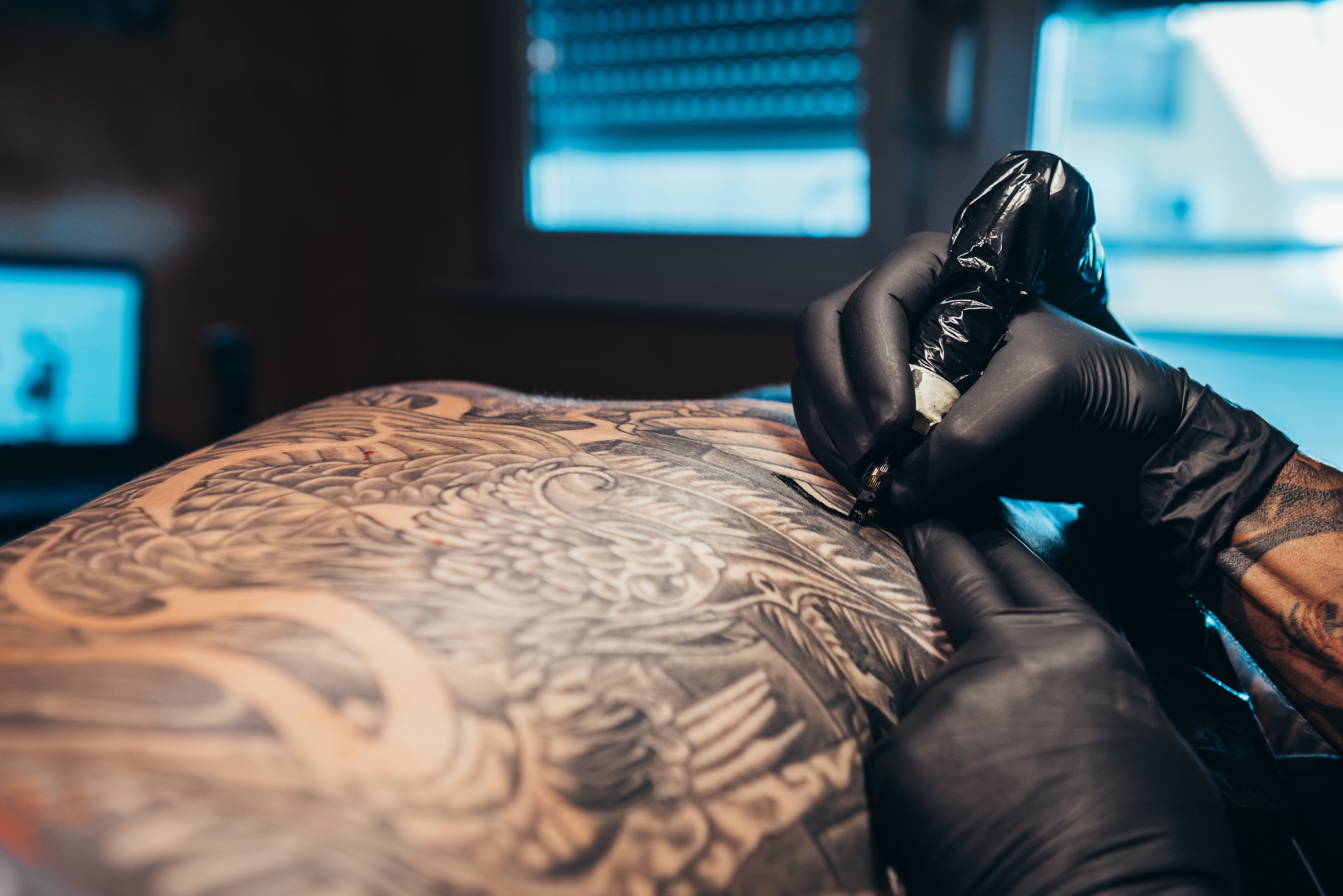 Hands of a tattoo artist wearing black gloves and holding a machine while creating a picture on a man back