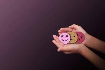 Hands holding three emotion face symbols sad, neutral, and happy on dark background representing mental health awareness, mood change, emotional balance, and psychological wellbeing,