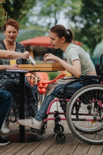 A boy with Down syndrome, a girl in a wheelchair, and an older woman play cards outside