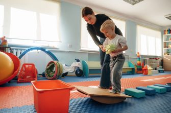 Teacher helping young child balance on a wobble board in a playroom