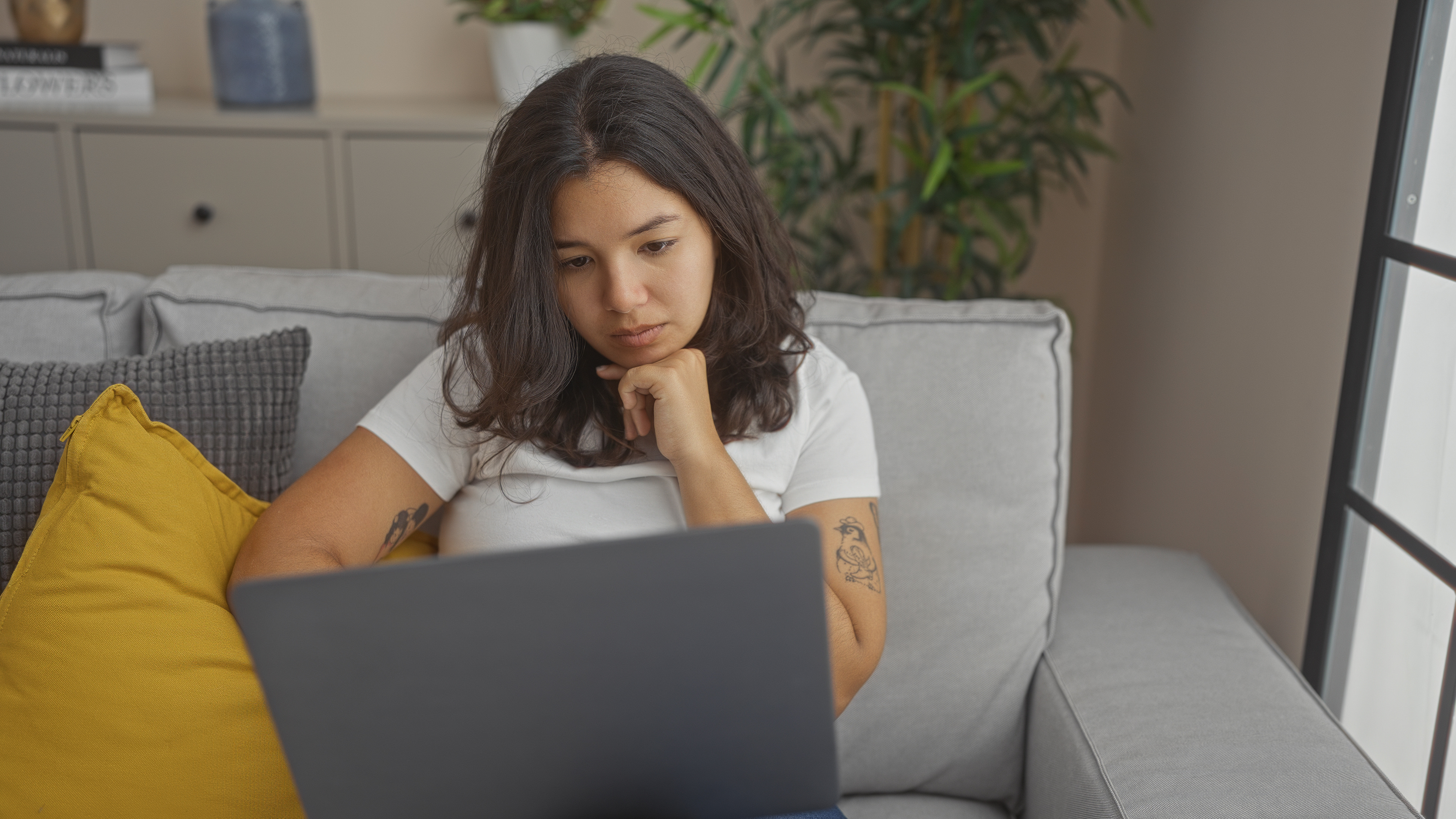 A young hispanic woman in a white shirt using a laptop while sitting on a gray sofa in a cozy living room.