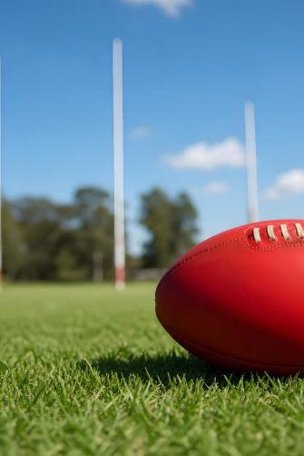 a red afl football rests on green grass in front of three afl posts on a sunny day