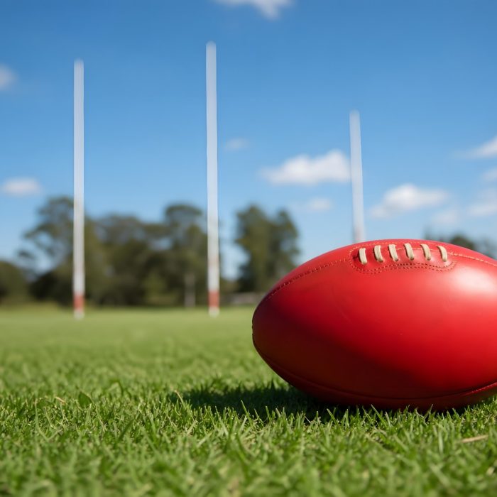 a red afl football rests on green grass in front of three afl posts on a sunny day