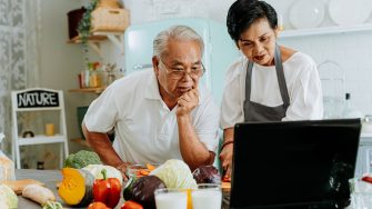 Senior Asian couple using a laptop while cooking in kitchen at home. 70s Elderly man and woman in relationship retirement lifestyle concept