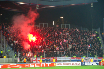 A soccer crowd behind high fences, watched by security as a flare is ignited.