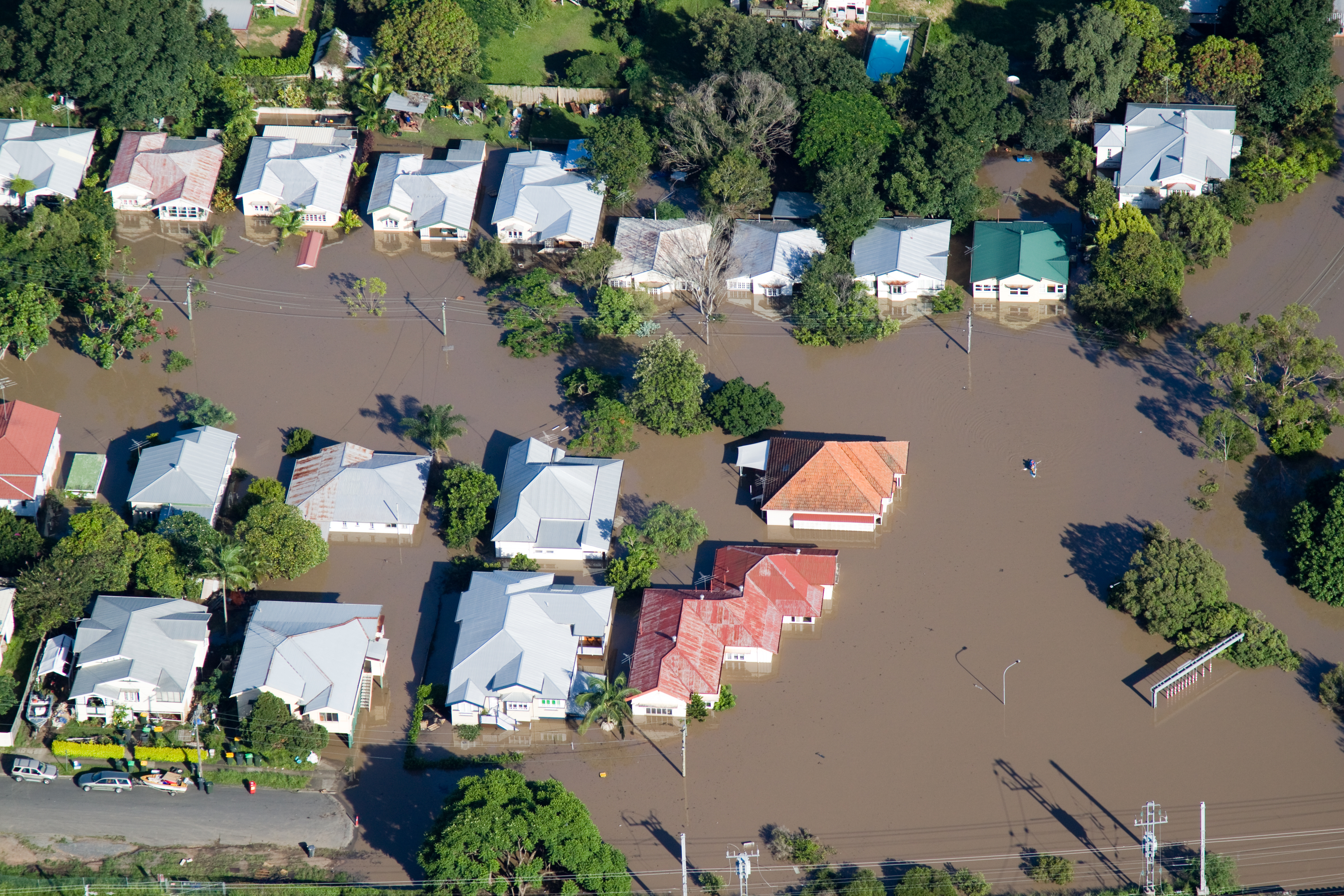 Muddy flood water surrounds a group of houses, with water almost up to the roof and roads completely covered