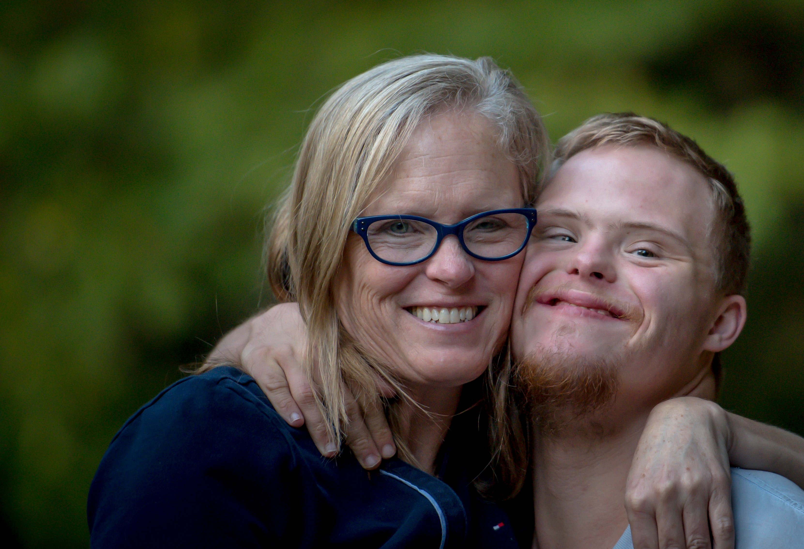 Two people share a close, smiling embrace in an outdoor setting with a blurred green background.