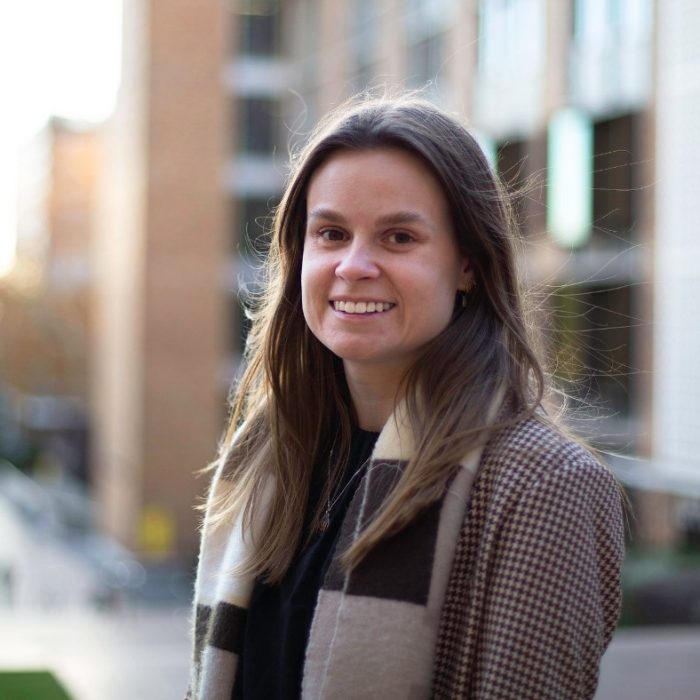 Young woman (Emily) smiling at the camera in May 2025. She is standing at the top of University Walk on the UNSW Sydney campus, hair blowing in the wind. The campus extends far behind her