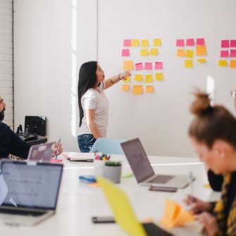 Young woman working with coloured sticky notes on board while colleagues look on from large meeting table