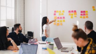 Young woman working with coloured sticky notes on board while colleagues look on from large meeting table