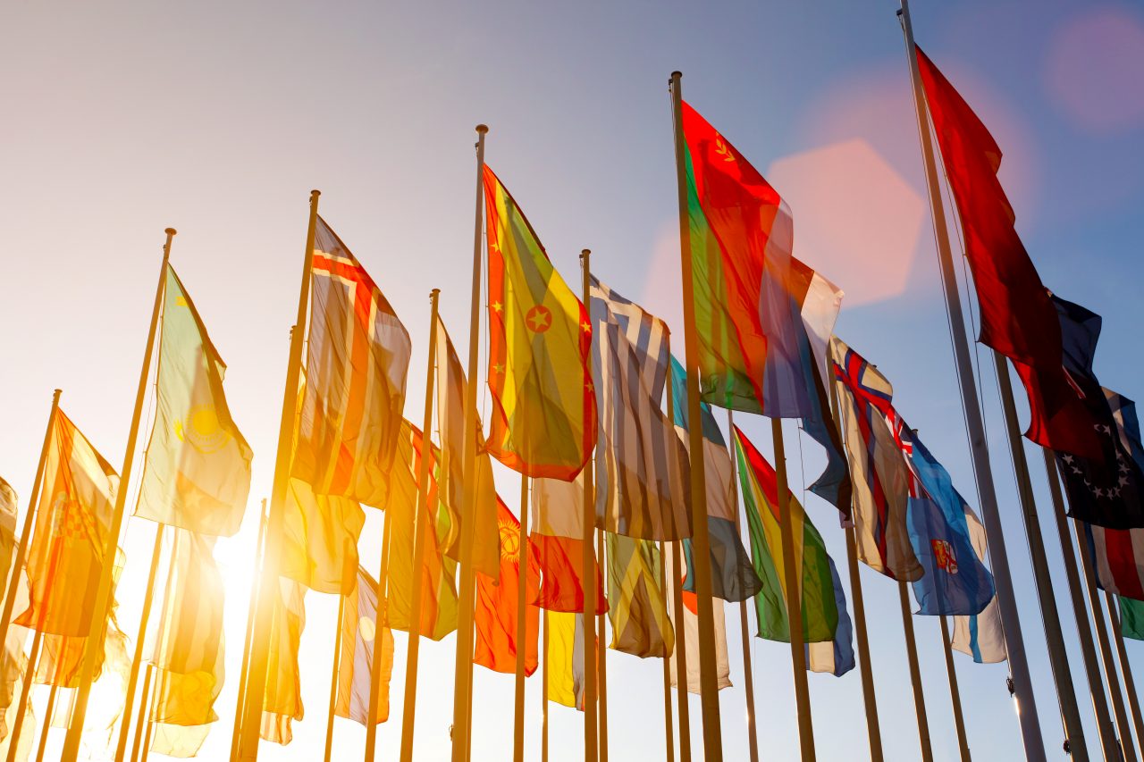 Group of many colorful internationl world flags waving in the sky at sunset, with shining sun and lens flare, low angle view.