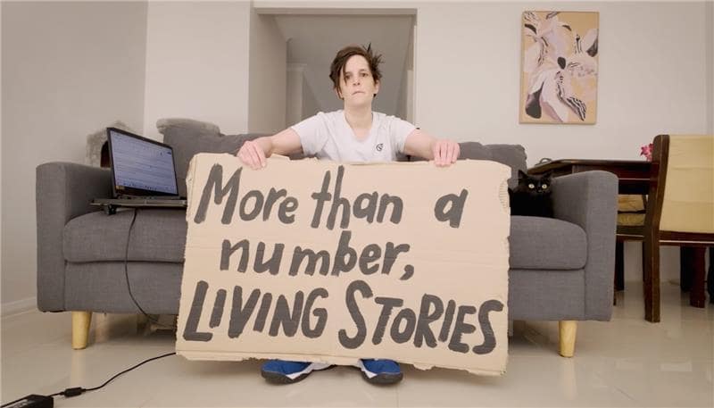 A young woman with short dark hair, a white t-shirt and sneakers sits on a grey couch holding a handwritten cardboard sign that reads “more than a number, living stories”. She is looking with a straight face directly at the camera, with a black cat sitting to one side of her and a laptop on the other