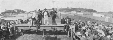 Black and white image of people attending an outdoor public meeting with four speakers on a wooden platform in 1919