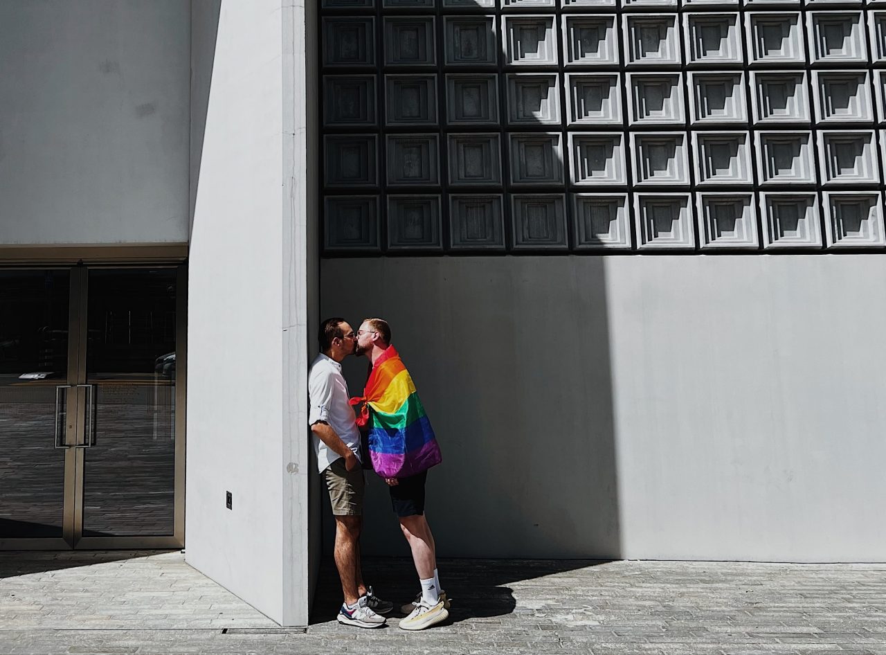 Two people kissing against building with one wrapped in gay pride flag