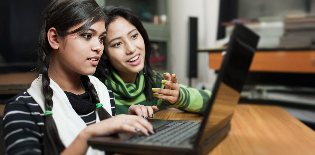 Indoor image of two late teen girls from different ethnicity using laptop together. Selective focus is on Indian girl sitting in foreground and typing on the keyboard while looking at laptop screen. Two people, waist up, horizontal composition with selective focus.