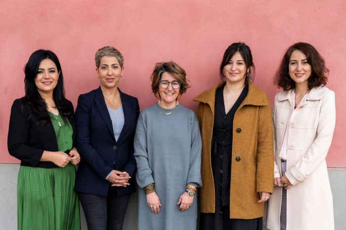Five smiling women standing against a pink wall background