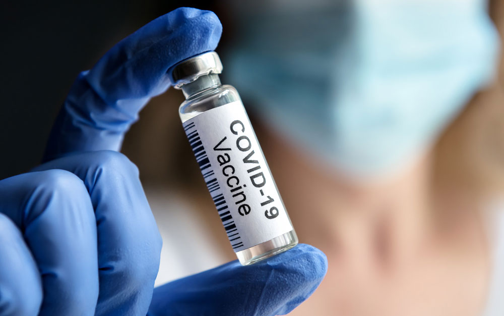 Gloved hand holding a vial which says "COVID-19 Vaccine"
