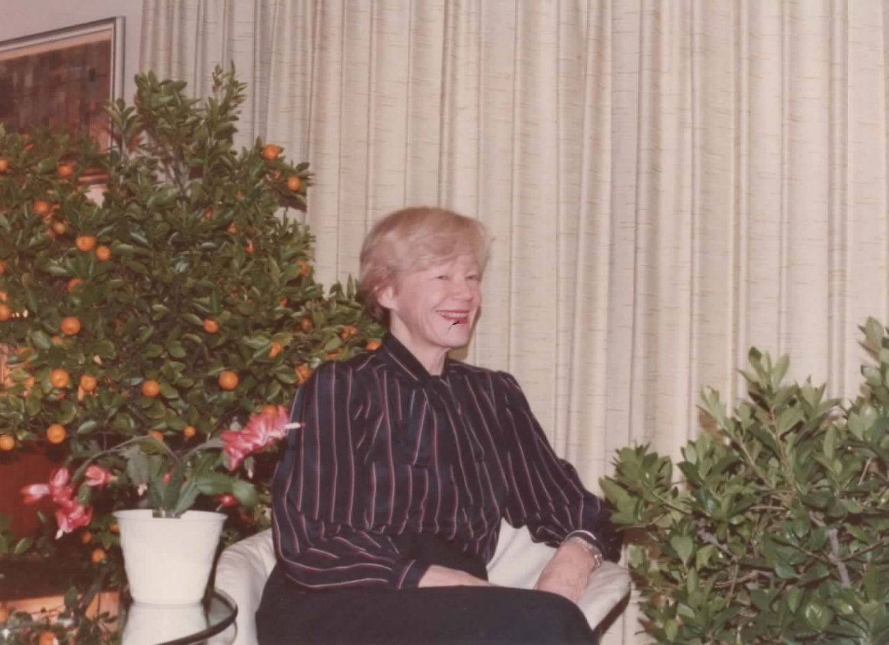A photo of Dr Helen Maguire sitting  in front of a curtain, surrounded by plants.