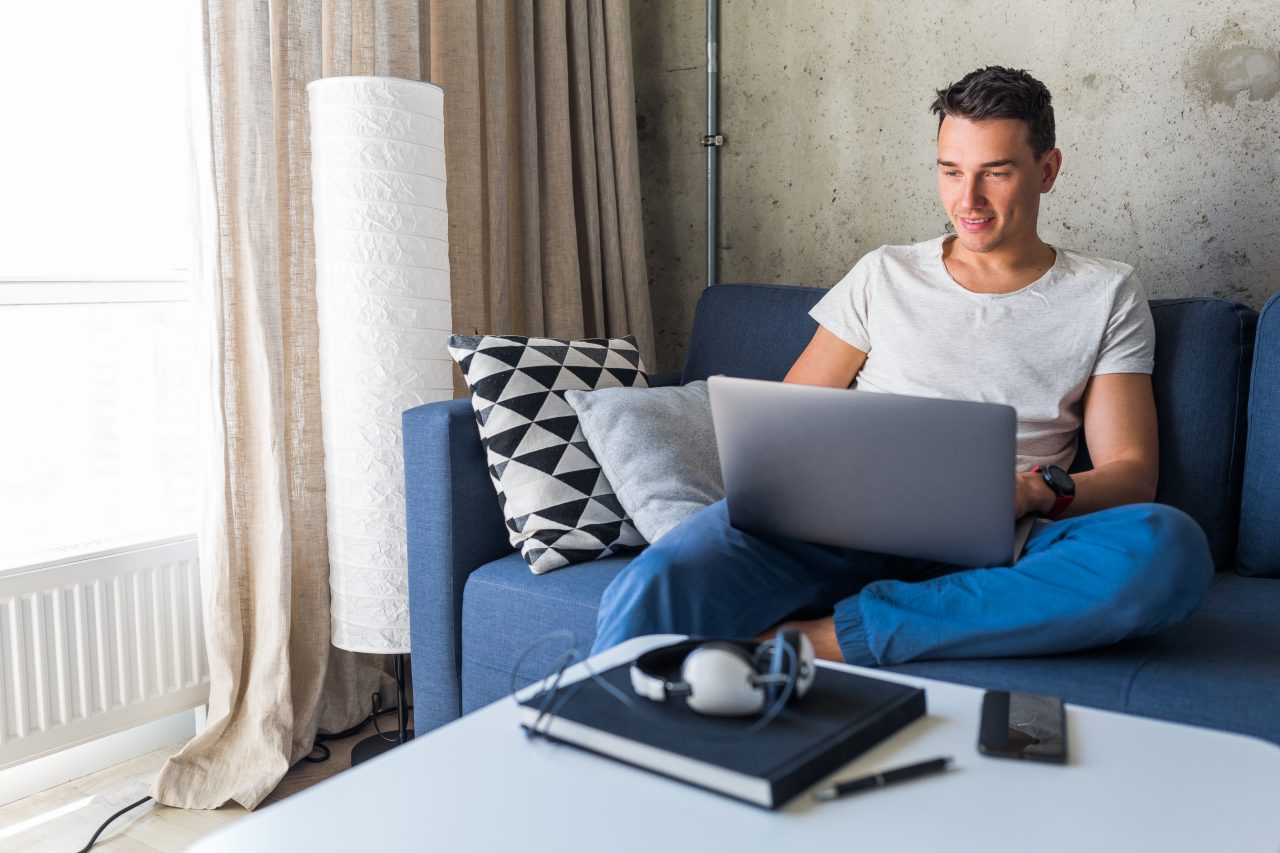 young attractive man sitting on sofa at home working on laptop online, using internet, smiling, happy mood, freelancer, free leisure time, relaxed, modern job lifestyle
