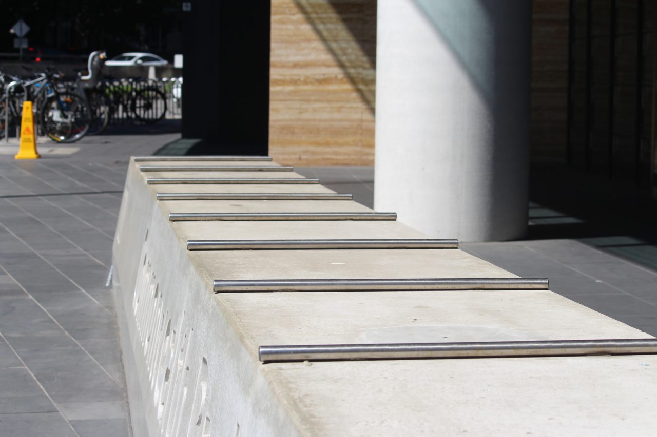 Concrete barrier / bench seat with fixed metal rods that prevent anyone from lying down on it (hostile design). The block of concrete has corporate signage on the front.