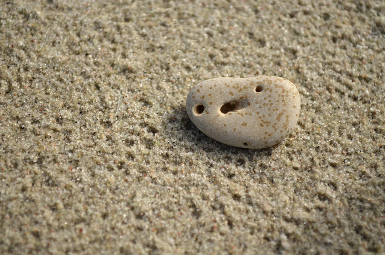 Pebble on sand, beach background. Funny stone with face, formed by holes. Illustration of pareidolia, optical illusion.