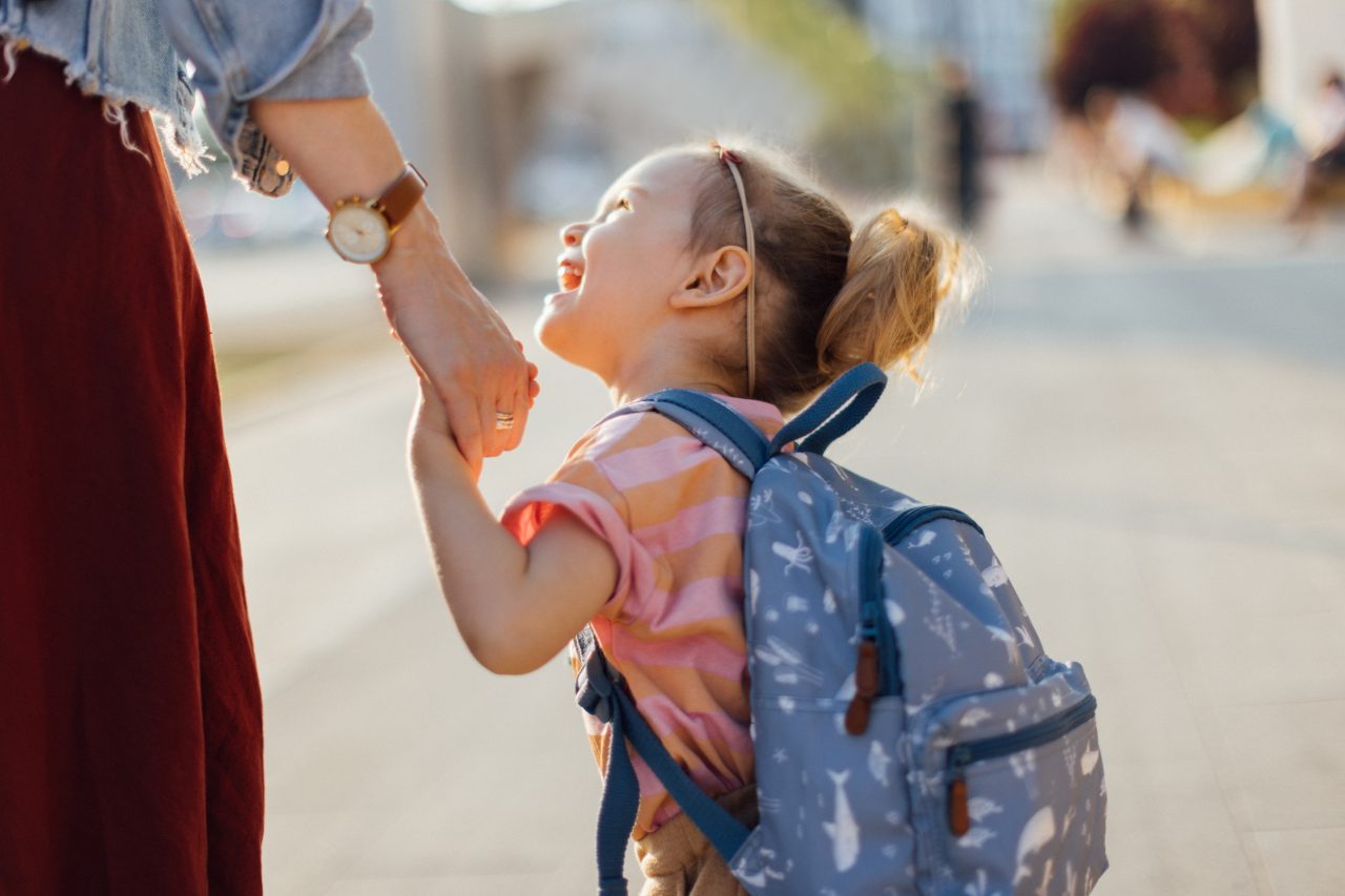 Smiling little girl with backpack holding her mum's hand while talking and walking to the school